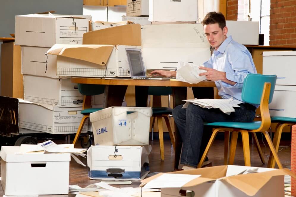 Man sitting at a desk sorting through piles and boxes of documents